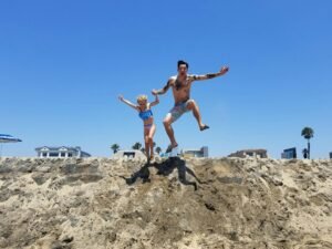 A father and daughter joyfully jumping on a sandy beach under a clear blue sky.